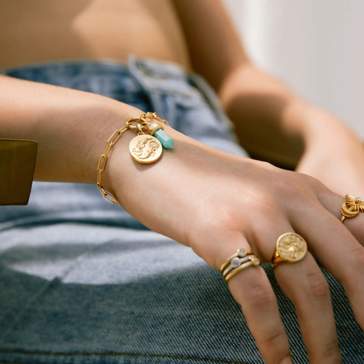 A person wearing a Single Charm Collector Bracelet by Awe Inspired adorned with gold charms and multiple rings on their fingers, resting their hand on their lap, dressed in denim.