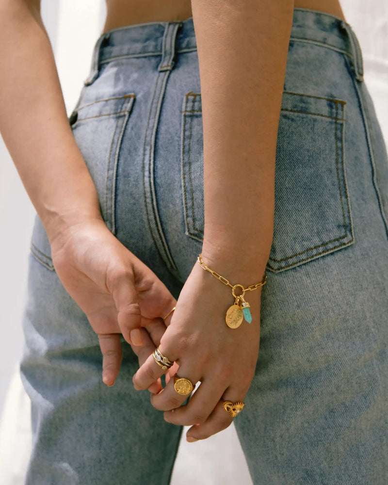 A person wearing blue jeans and various gold rings stands with hands behind their back, their Single Charm Collector Bracelet by Awe Inspired glinting alongside other jewelry.
