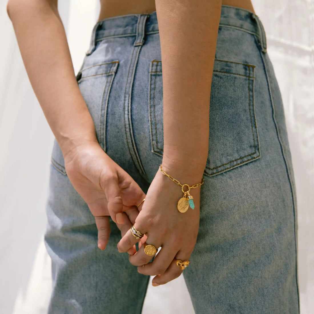 A person wearing blue jeans and various gold rings stands with hands behind their back, their Single Charm Collector Bracelet by Awe Inspired glinting alongside other jewelry.