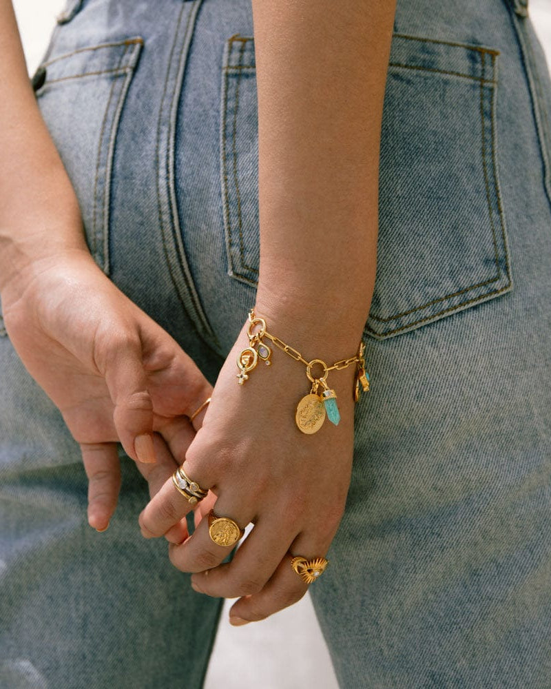 A person wearing multiple rings and The Charm Collector Bracelet by Awe Inspired rests their hands behind their back. The bracelet, capturing a 90s charm bracelet aesthetic, adorns the individual's wrist as they stand casually in blue jeans.
