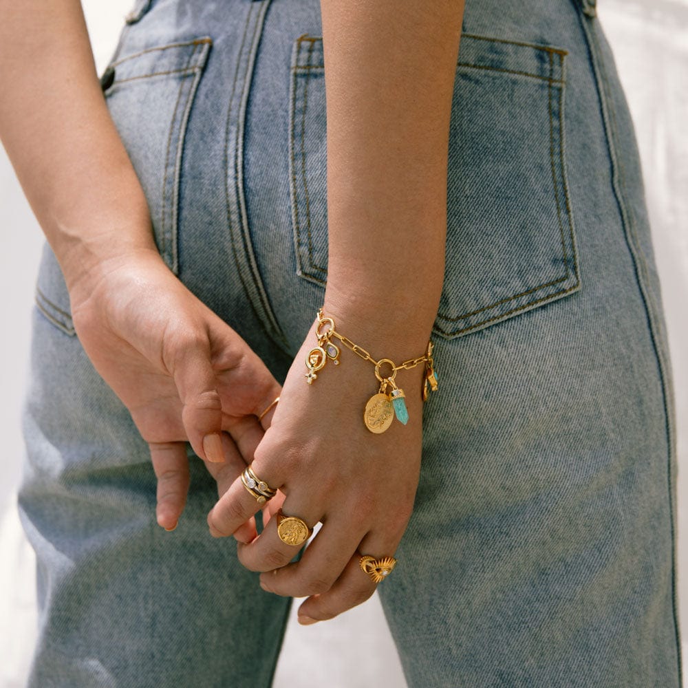 A person wearing multiple rings and The Charm Collector Bracelet by Awe Inspired rests their hands behind their back. The bracelet, capturing a 90s charm bracelet aesthetic, adorns the individual's wrist as they stand casually in blue jeans.