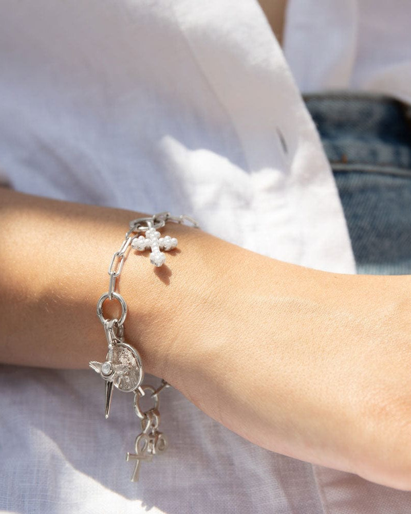 Close-up of a person wearing The Charm Collector Bracelet by Awe Inspired, adorned with a cross, heart, and various small charms, capturing the 90s charm bracelet aesthetic. The background reveals part of a white shirt and denim jeans.