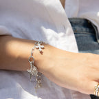 Close-up of a person wearing The Charm Collector Bracelet by Awe Inspired, adorned with a cross, heart, and various small charms, capturing the 90s charm bracelet aesthetic. The background reveals part of a white shirt and denim jeans.