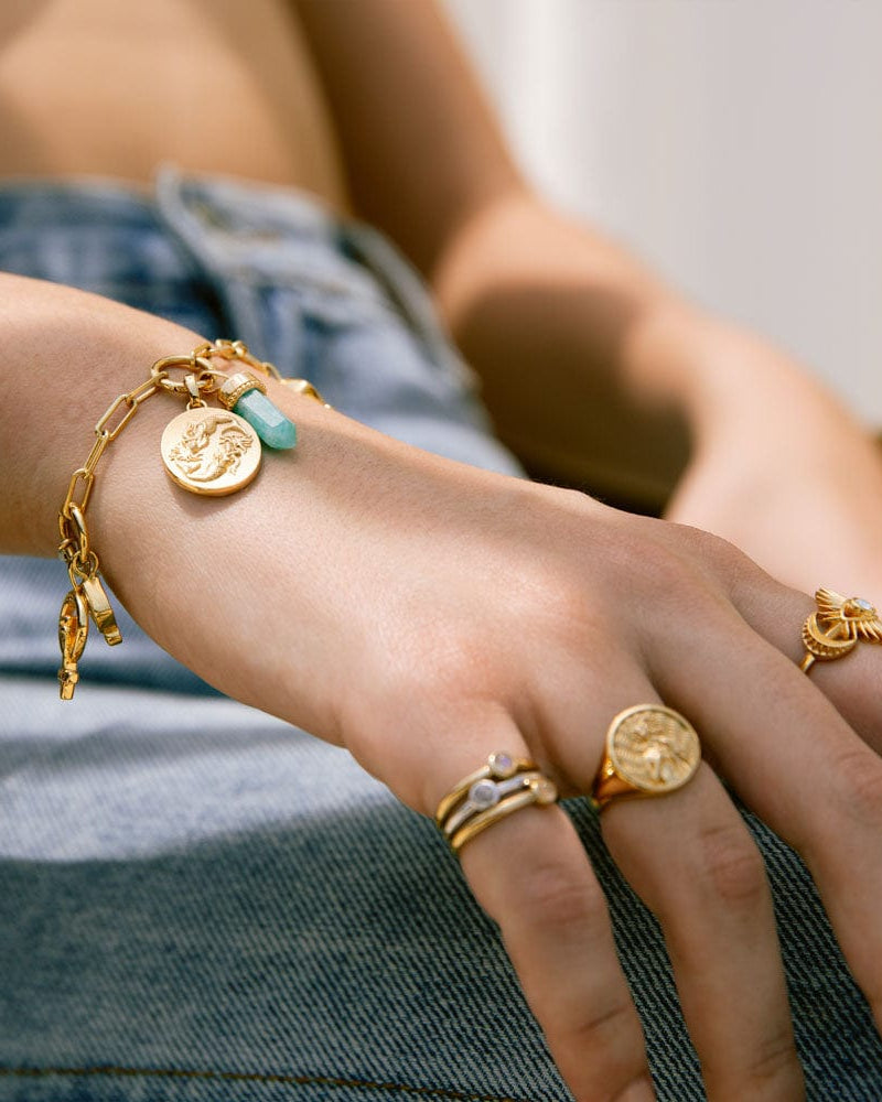 A hand adorned with gold jewelry, including Awe Inspired’s The Charm Collector Bracelet featuring a 90s aesthetic with coins and turquoise stone, plus several rings, rests on a denim-clad leg.