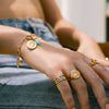 A hand adorned with gold jewelry, including Awe Inspired’s The Charm Collector Bracelet featuring a 90s aesthetic with coins and turquoise stone, plus several rings, rests on a denim-clad leg.