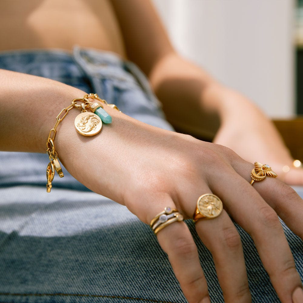 A hand adorned with gold jewelry, including Awe Inspired’s The Charm Collector Bracelet featuring a 90s aesthetic with coins and turquoise stone, plus several rings, rests on a denim-clad leg.