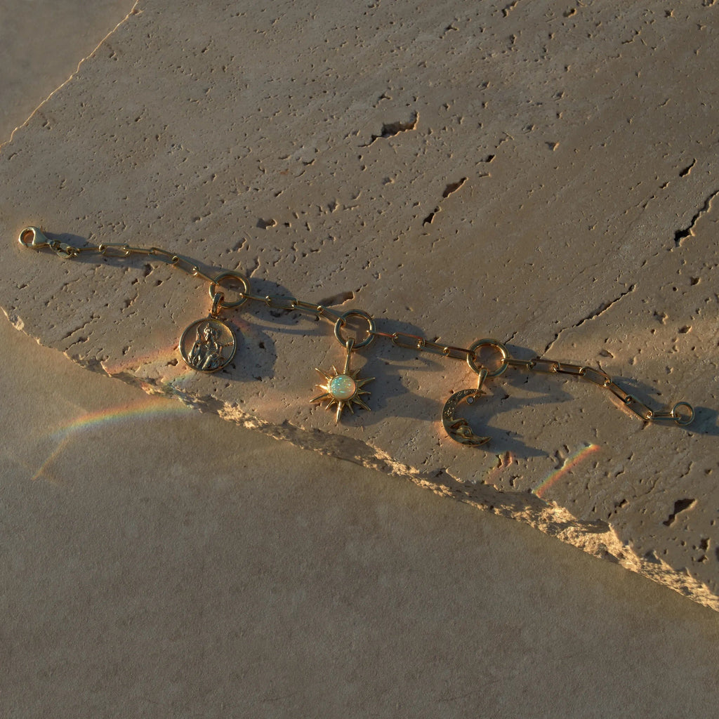 The Charm Collector Bracelet by Awe Inspired, crafted with a gold chain and adorned with three charms—an iconic figure, a sun set with a white gem, and a crescent moon—rests elegantly on a textured stone surface, its light reflections capturing the nostalgic 90s charm bracelet aesthetic.