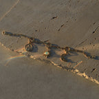 The Charm Collector Bracelet by Awe Inspired, crafted with a gold chain and adorned with three charms—an iconic figure, a sun set with a white gem, and a crescent moon—rests elegantly on a textured stone surface, its light reflections capturing the nostalgic 90s charm bracelet aesthetic.