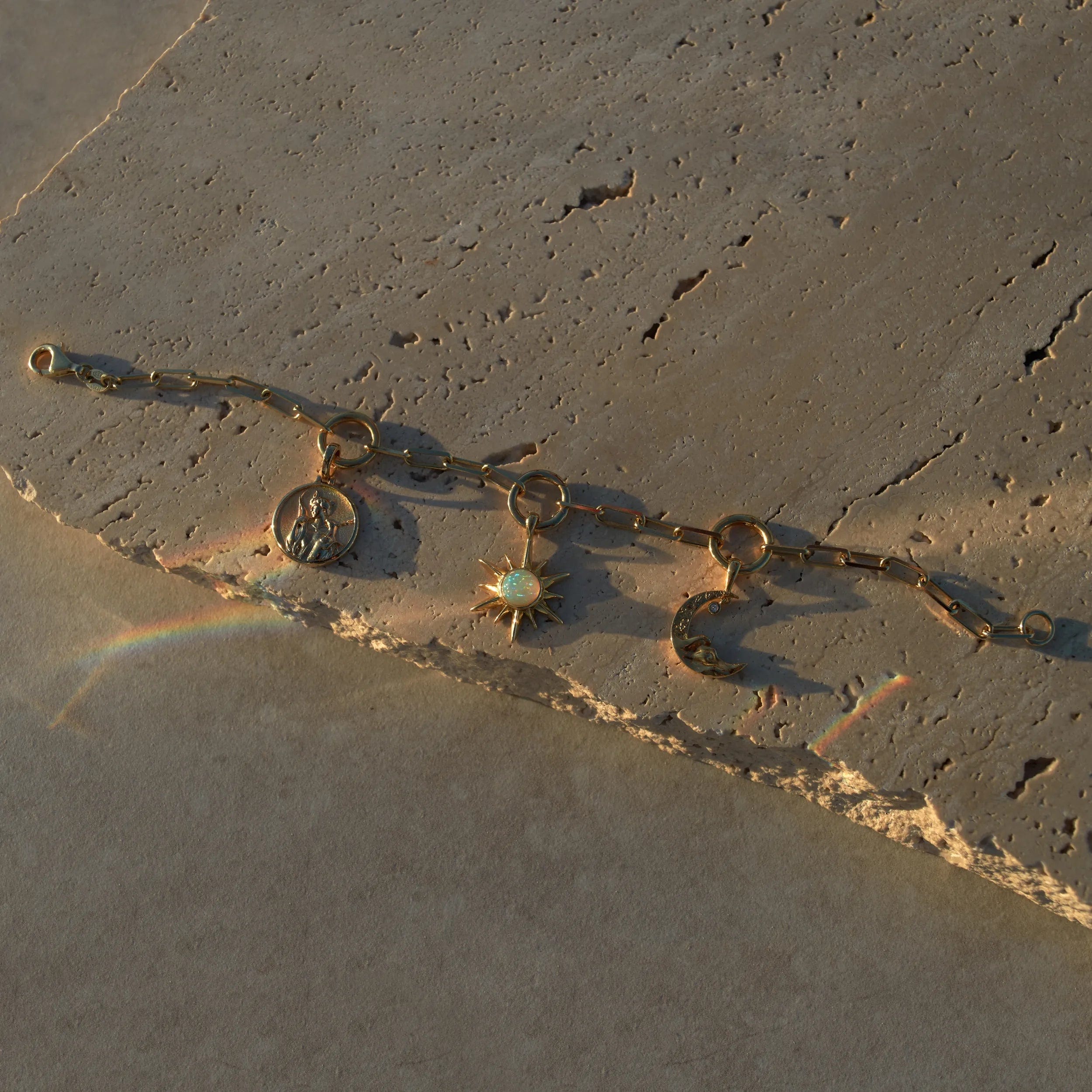 The Charm Collector Bracelet by Awe Inspired, crafted with a gold chain and adorned with three charms—an iconic figure, a sun set with a white gem, and a crescent moon—rests elegantly on a textured stone surface, its light reflections capturing the nostalgic 90s charm bracelet aesthetic.