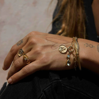 A hand adorned with intricate tattoos wears various pieces of gold jewelry, including the Wild Goddess Charm Bracelet by Awe Inspired, along with rings and bracelets featuring bezel set gemstones and charms. Like a talisman from the Goddess of the wilds, these treasures rest gracefully on a dark fabric background, exuding an aura of mystique.