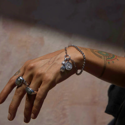 A tattoo-adorned hand, adorned with rings and the Awe Inspired Wild Goddess Charm Bracelet featuring bezel-set gemstones and charms, stands out against a softly lit background.