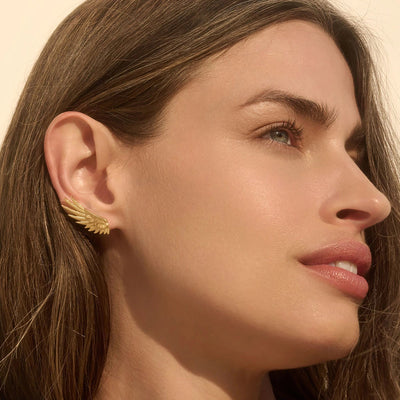 A close-up of a person's face with long hair, wearing a gold earring shaped like a wing, resembling the Awe Inspired Winged Ear Crawler. Their gaze is directed upwards, and the background is neutral.