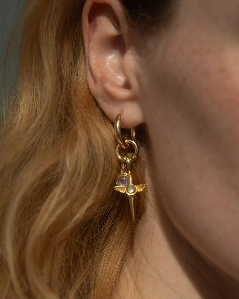 Close-up of a person wearing the Blood Moon Rising Charm Earrings by Awe Inspired, featuring a gold earring with a small dangling charm shaped like a sword adorned with a purple gemstone. The person's light brown, wavy hair is partially shown—a true delight for any Charm Collector.