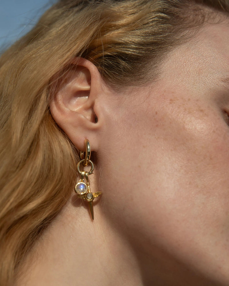 Close-up of a person with light hair wearing the "Blood Moon Rising Charm Earrings" by Awe Inspired, which feature a dangling design with a Rainbow Moonstone amulet and a small spike.