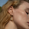 Close-up of a person with light hair wearing the "Blood Moon Rising Charm Earrings" by Awe Inspired, which feature a dangling design with a Rainbow Moonstone amulet and a small spike.