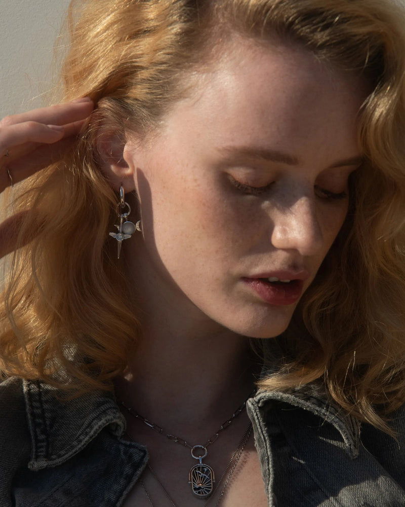 A person with wavy blonde hair is touching their ear and wearing a denim jacket, Awe Inspired Blood Moon Rising Charm Earrings, and a silver necklace with a circular pendant.