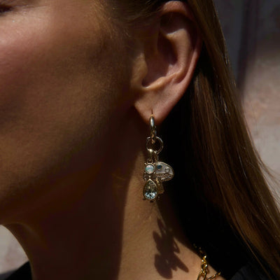 Close-up of a woman's ear wearing the Awe Inspired Magic Mushroom Single Charm Earring, which features a gold hoop adorned with a Magic Mushroom charm, a small gemstone talisman, and a tear-drop shaped pendant. The background is blurred.