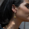 Close-up side profile of a person wearing the Natural Aura Charm Earrings by Awe Inspired, featuring multiple dangling charms adorned with small stones, geometric shapes, and a glistening green amethyst.