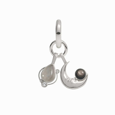 The "In My Orbit Charm Earring" by Awe Inspired features a teardrop-shaped silver pendant adorned with small crystal accents on the left and a crescent moon design with a dark spherical stone on the right, all connected by a loop, embodying the essence of an Astral Amulet.