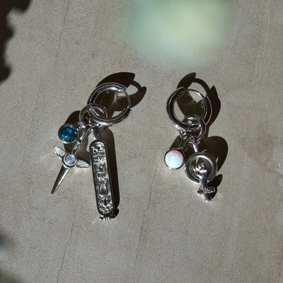 Two keychains with multiple charms: the left one features a turquoise gem, cross with wings, and an engraved bar from The Charm Collector Earrings collection by Awe Inspired in silver, while the right one showcases a round opal, globe and ring, and a female symbol from the same stunning collection.