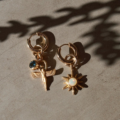 A pair of 'The Charm Collector Earrings' by Awe Inspired rests on a stone surface. One earring features a dangling figure with wings and a blue gem, while the other boasts a sun pendant with an opal center. Shadows fall across these Awe Inspired charms, creating an air of mystery and allure.