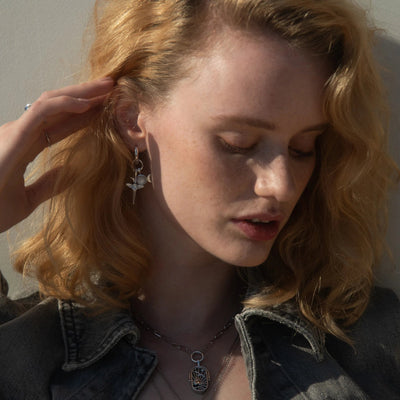 A person with wavy blonde hair, wearing a denim jacket, a necklace with a large pendant, and The Charm Collector Earrings by Awe Inspired, tilts their head down and touches their hair.