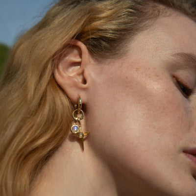 Close-up of a person's ear adorned with "The Charm Collector Earrings" by Awe Inspired, featuring multiple charms including a small jewel and a spike. The person has light skin and blonde hair.