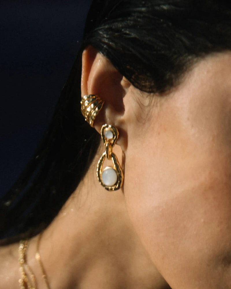 A close-up of a person's ear adorned with gold earrings, one featuring a pear-shaped White Topaz, the gemstone of manifestation. The person also wears an elegant gold ear cuff called the Topaz Claw Ear Cuff by Awe Inspired and has dark hair cascading next to a matching gold necklace.