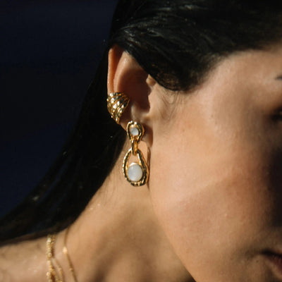 A close-up of a person's ear adorned with gold earrings, one featuring a pear-shaped White Topaz, the gemstone of manifestation. The person also wears an elegant gold ear cuff called the Topaz Claw Ear Cuff by Awe Inspired and has dark hair cascading next to a matching gold necklace.