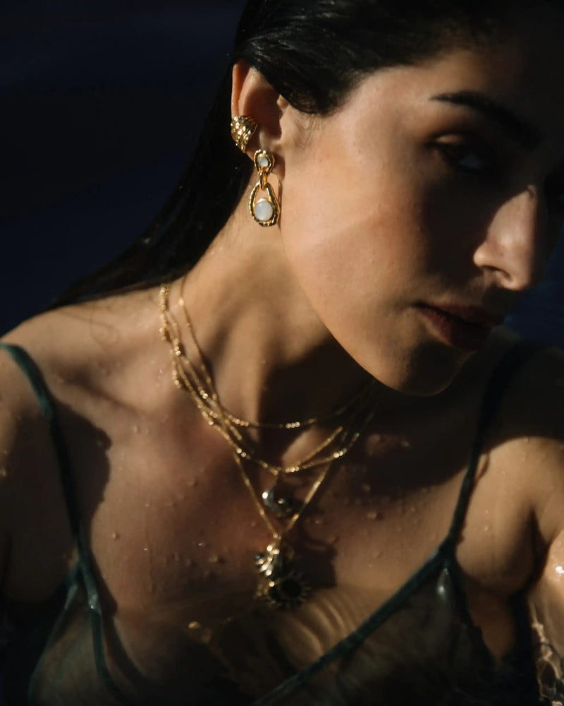 A woman with wet hair wears gold jewelry, including an Awe Inspired Topaz Claw Ear Cuff and layered necklaces with White Topaz gemstones. Partially submerged in dark water, her face and upper body are illuminated, creating a striking contrast with the background.