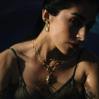 A woman with wet hair wears gold jewelry, including an Awe Inspired Topaz Claw Ear Cuff and layered necklaces with White Topaz gemstones. Partially submerged in dark water, her face and upper body are illuminated, creating a striking contrast with the background.