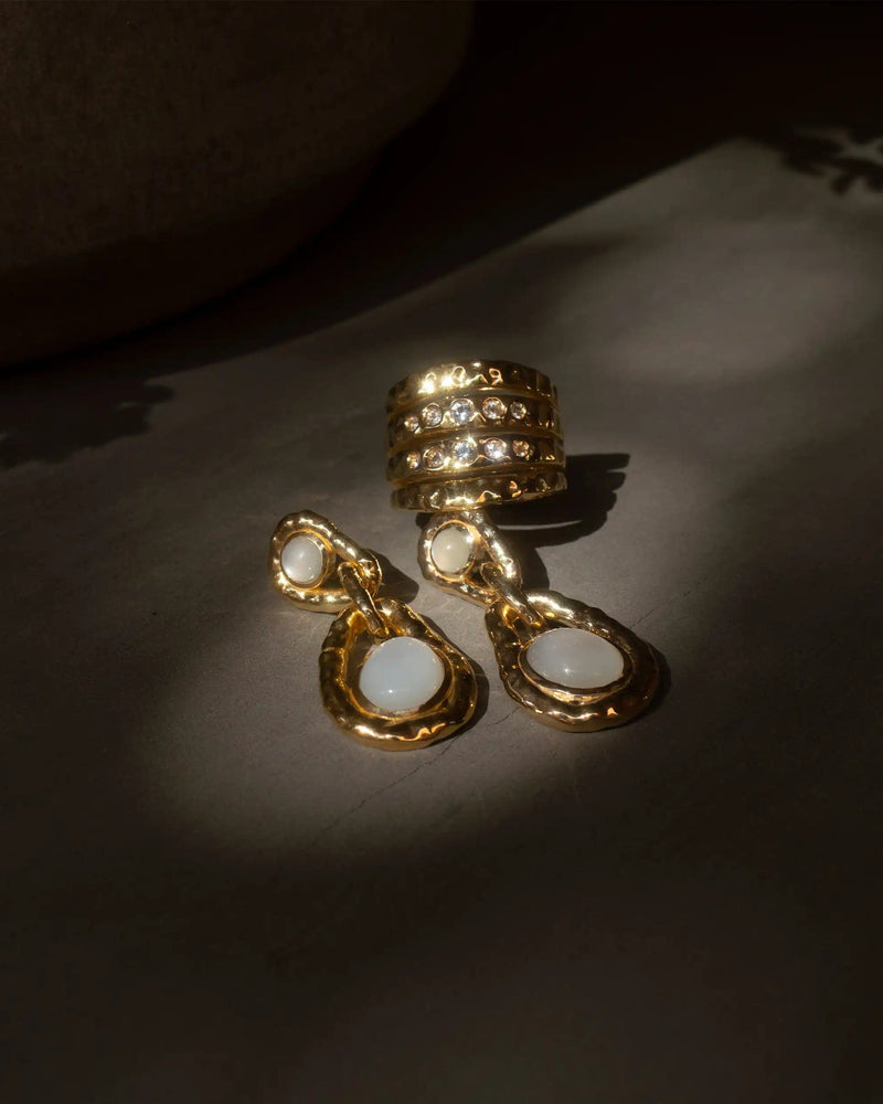 A gold ring with multiple cubic zirconia stones, a pair of gold dangle earrings with white stones, and a delicate Topaz Claw Ear Cuff by Awe Inspired featuring White Topaz gemstones are displayed on a dark surface.