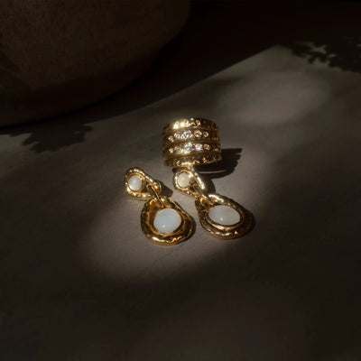 A gold ring with multiple cubic zirconia stones, a pair of gold dangle earrings with white stones, and a delicate Topaz Claw Ear Cuff by Awe Inspired featuring White Topaz gemstones are displayed on a dark surface.