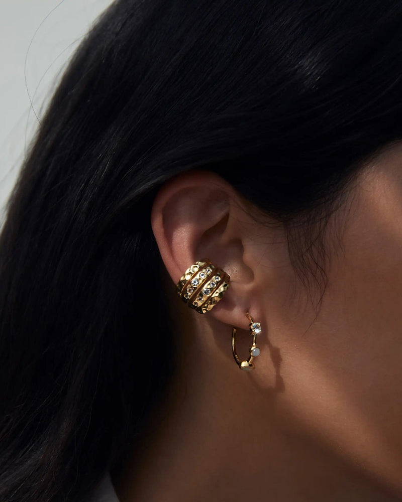 Close-up of a person's ear adorned with two earrings: one is a gold hoop with small charms, and the other is an upper ear cuff featuring an intricate design highlighted by a sparkling White Topaz gemstone from the Topaz Claw Ear Cuff by Awe Inspired. Dark hair partially covers the ear.