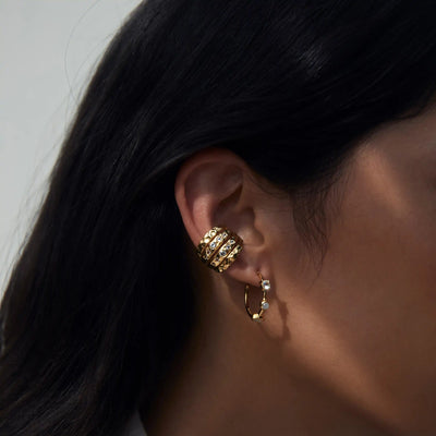 Close-up of a person's ear adorned with two earrings: one is a gold hoop with small charms, and the other is an upper ear cuff featuring an intricate design highlighted by a sparkling White Topaz gemstone from the Topaz Claw Ear Cuff by Awe Inspired. Dark hair partially covers the ear.