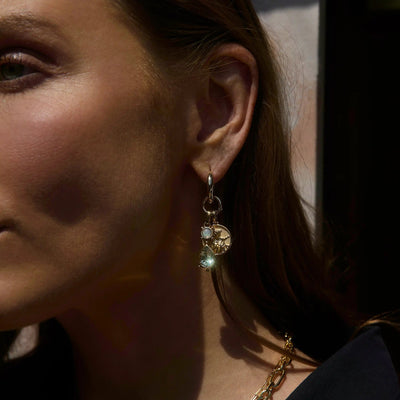 Close-up of a woman wearing the Wild Goddess Single Charm Earring by Awe Inspired, featuring multiple charms including a gemstone talisman and a circular pendant. Only the right side of her face is visible, showing part of her ear and cheek, evoking an air as wild and mysterious as Artemis herself.