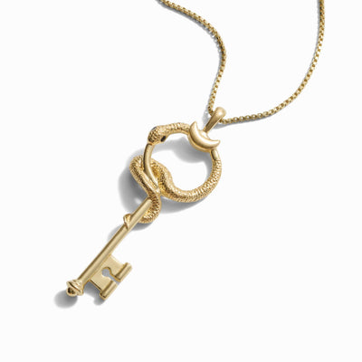 A gold necklace featuring a key pendant entwined with a snake design on a thin chain, isolated on a white background, reminiscent of The Gate of Hades Key Necklace by Awe Inspired, symbolizing hidden power.