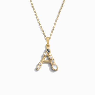 The Gold Gemstone Initial Necklace by Awe Inspired features a gold chain adorned with a pendant shaped like the letter "A," embellished with bezel-set gemstones for extra brilliance. It's perfect for making a statement with its elegantly designed initial charm.