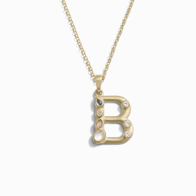 The Gold Gemstone Initial Necklace by Awe Inspired features a gold necklace adorned with a statement-sized initial charm in the shape of the letter "B," embellished with various small bezel-set gemstones.