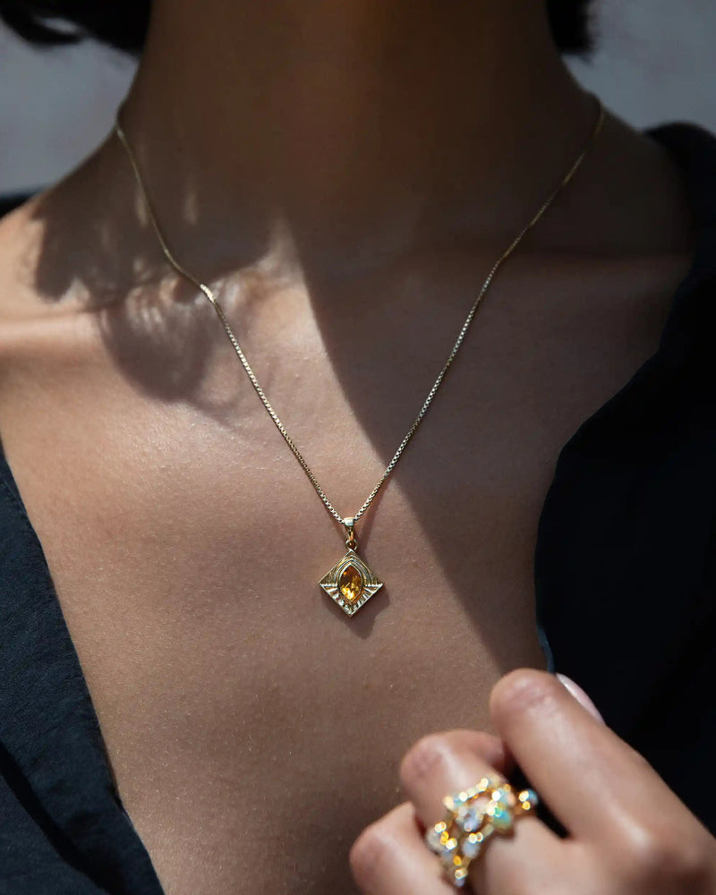 Close-up of a person wearing the Aura Pendant by Awe Inspired, exuding an aura of elegance, and multiple gold rings with gemstones. The person is dressed in a dark top.