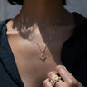 Close-up of a person wearing the Aura Pendant by Awe Inspired, exuding an aura of elegance, and multiple gold rings with gemstones. The person is dressed in a dark top.