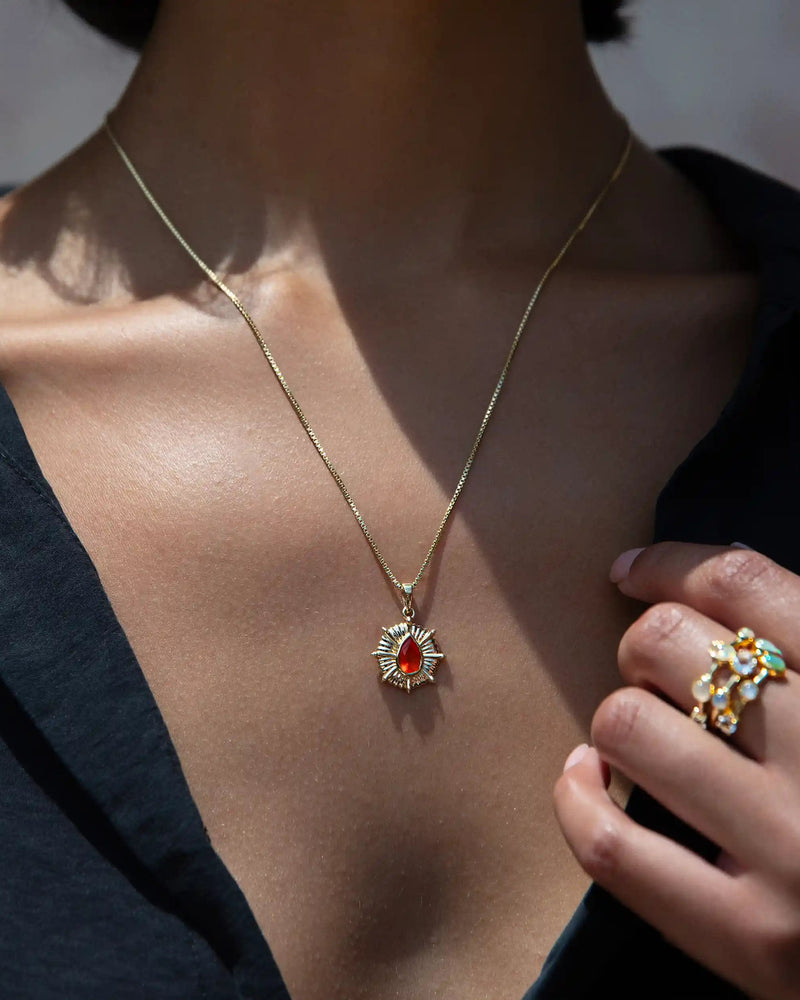 Close-up of a person wearing the Awe Inspired Aura Pendant, a gold necklace with a sun-shaped pendant featuring a red gemstone believed to balance the aura, and an ornate ring with colored gemstones on their hand.