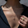 Close-up of a person wearing the Awe Inspired Aura Pendant, a gold necklace with a sun-shaped pendant featuring a red gemstone believed to balance the aura, and an ornate ring with colored gemstones on their hand.