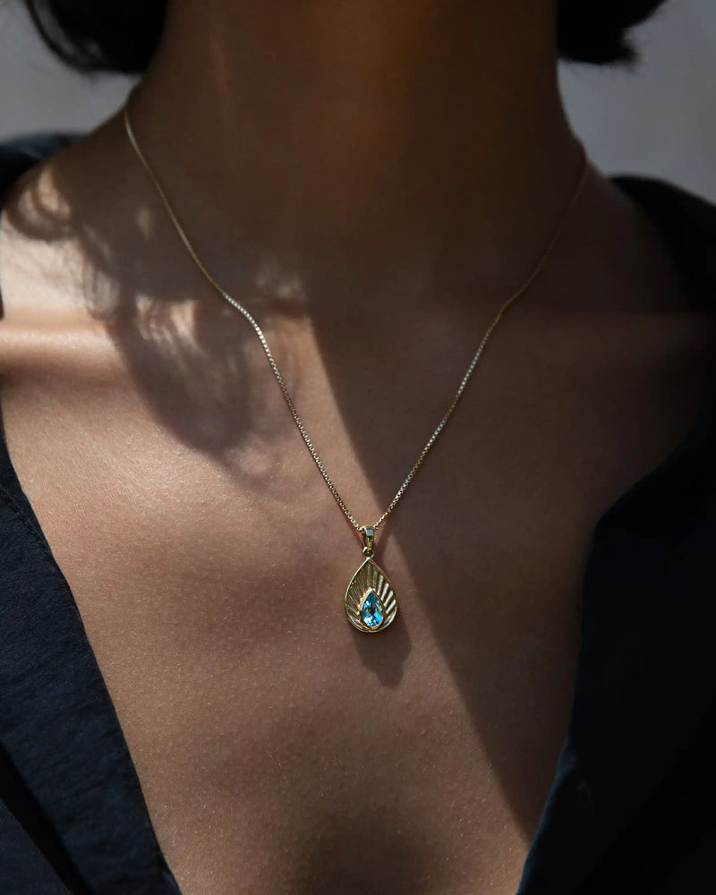 Close-up of a person wearing the Aura Pendant by Awe Inspired, a gold necklace with a teardrop-shaped pendant featuring a blue gemstone believed to harmonize the chakra, against a dark shirt.