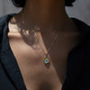 Close-up of a person wearing the Aura Pendant by Awe Inspired, a gold necklace with a teardrop-shaped pendant featuring a blue gemstone believed to harmonize the chakra, against a dark shirt.