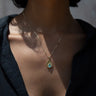 Close-up of a person wearing the Aura Pendant by Awe Inspired, a gold necklace with a teardrop-shaped pendant featuring a blue gemstone believed to harmonize the chakra, against a dark shirt.