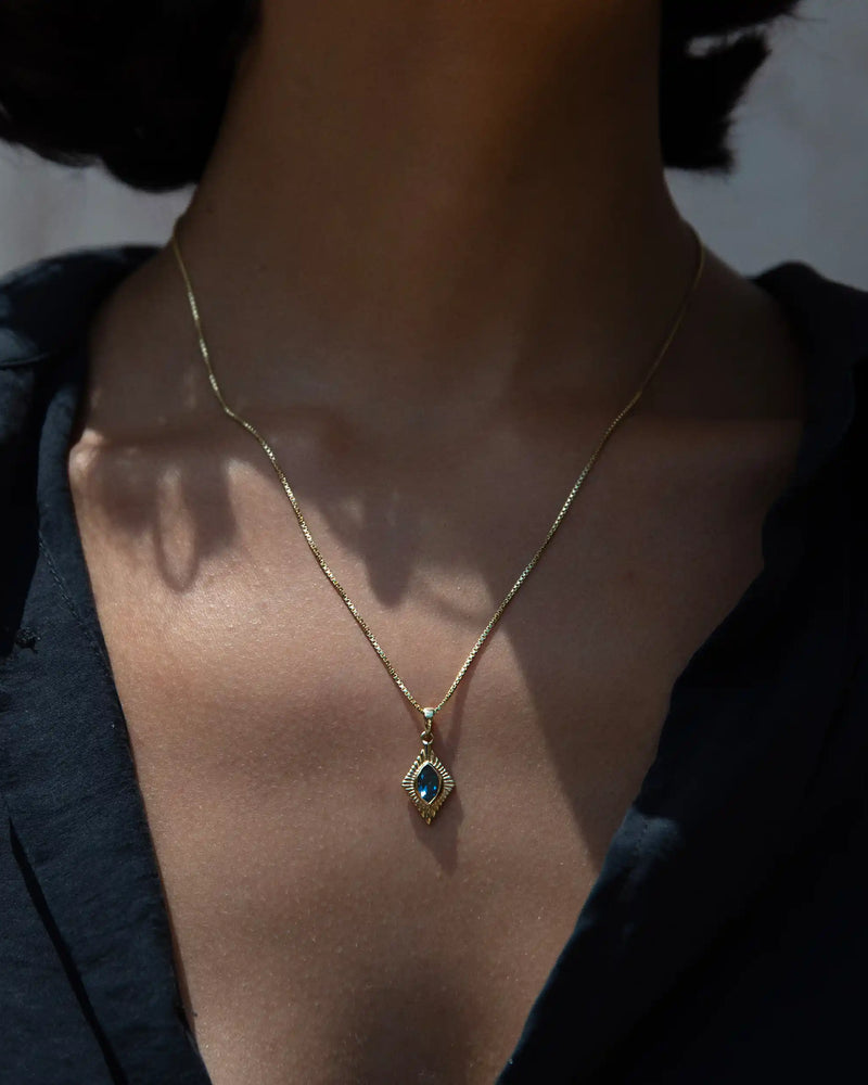 Close-up of a person wearing a black shirt and the Aura Pendant by Awe Inspired, featuring a gold necklace with a blue gemstone pendant, exuding an aura of elegance.
