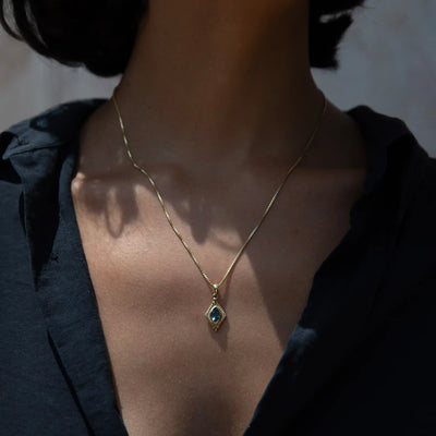 Close-up of a person wearing a black shirt and the Aura Pendant by Awe Inspired, featuring a gold necklace with a blue gemstone pendant, exuding an aura of elegance.