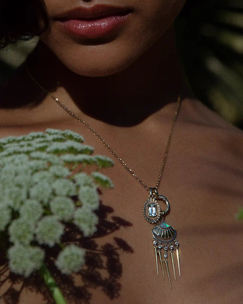 A woman adorned with the "Aura Pendant" by Awe Inspired, a gold necklace featuring a distinctive gemstone design, is partially obscured by soft-focus flowers in the foreground, giving her an ethereal aura.