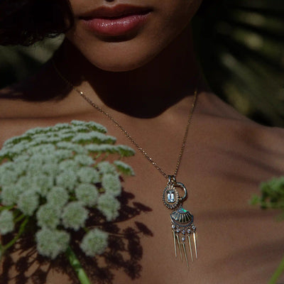 A woman adorned with the "Aura Pendant" by Awe Inspired, a gold necklace featuring a distinctive gemstone design, is partially obscured by soft-focus flowers in the foreground, giving her an ethereal aura.